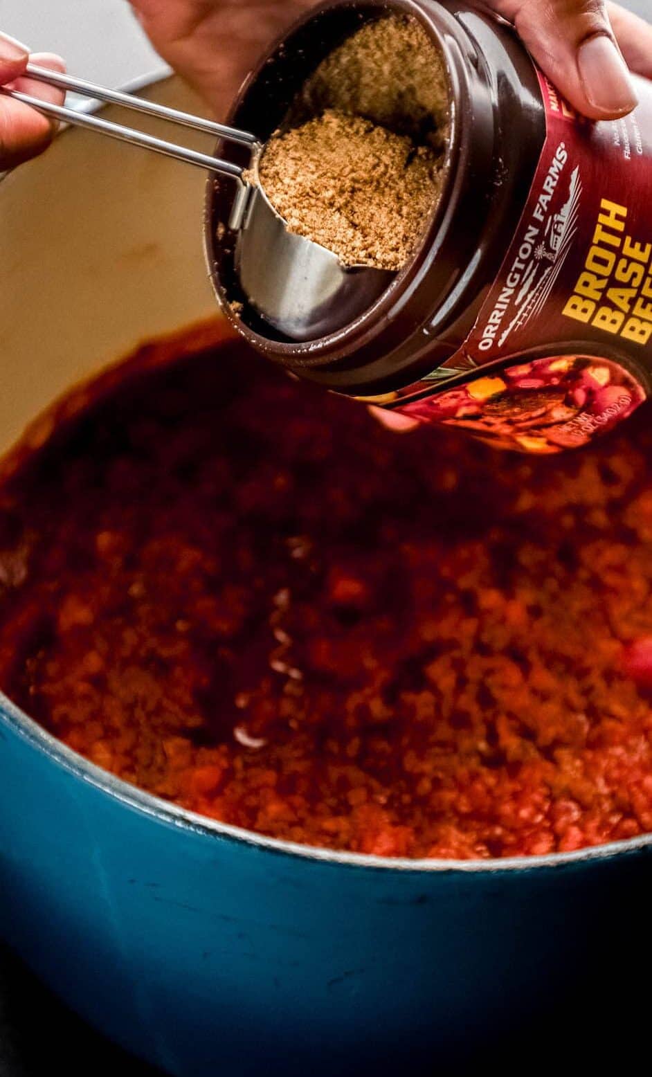 Hand scooping broth base mixture into pot with the other soup ingredients. 