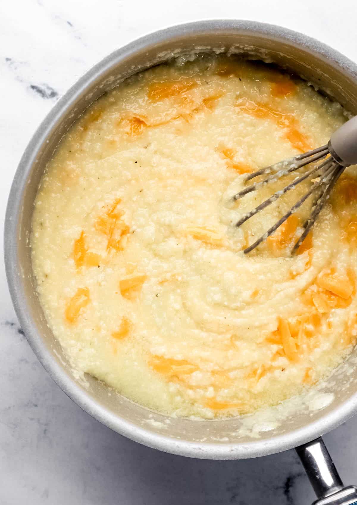 Shredded cheese and butter added to saucepan with girts and whisk in it. 