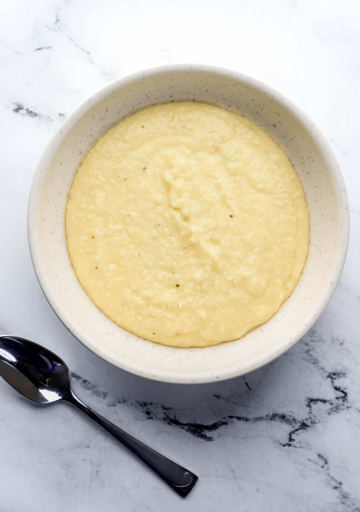 Overhead view of cheese grits in white bowl by a spoon on marble surface. 