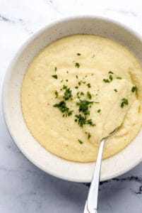 Overhead view of bowl of cheese grits topped with fresh parsley with spoon in it on marble surface.