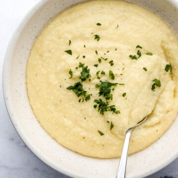 Overhead view of bowl of cheese grits topped with fresh parsley with spoon in it on marble surface.