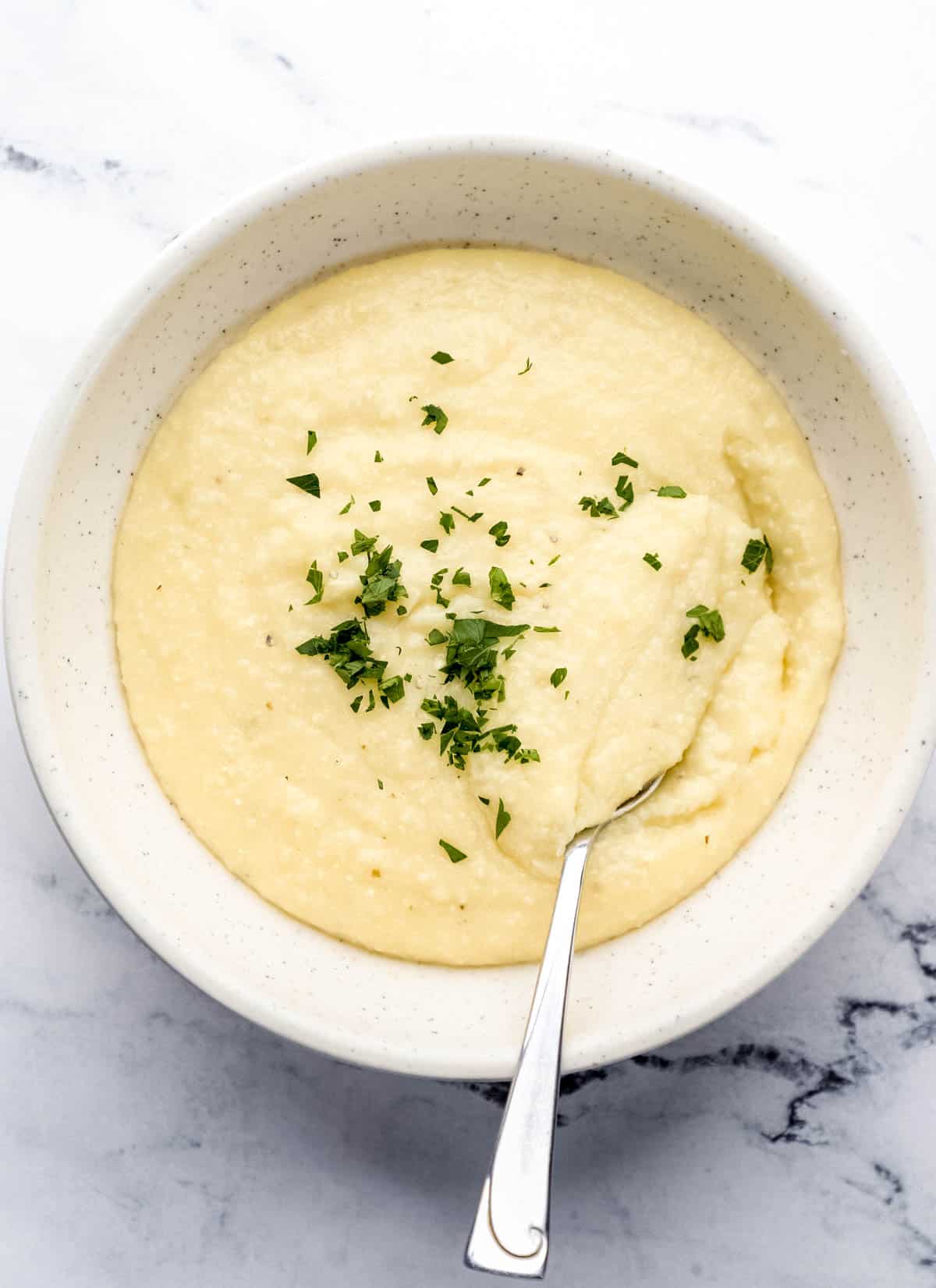 Overhead view of white bowl of cheese grits topped with fresh chopped parsley with a spoon in it. 