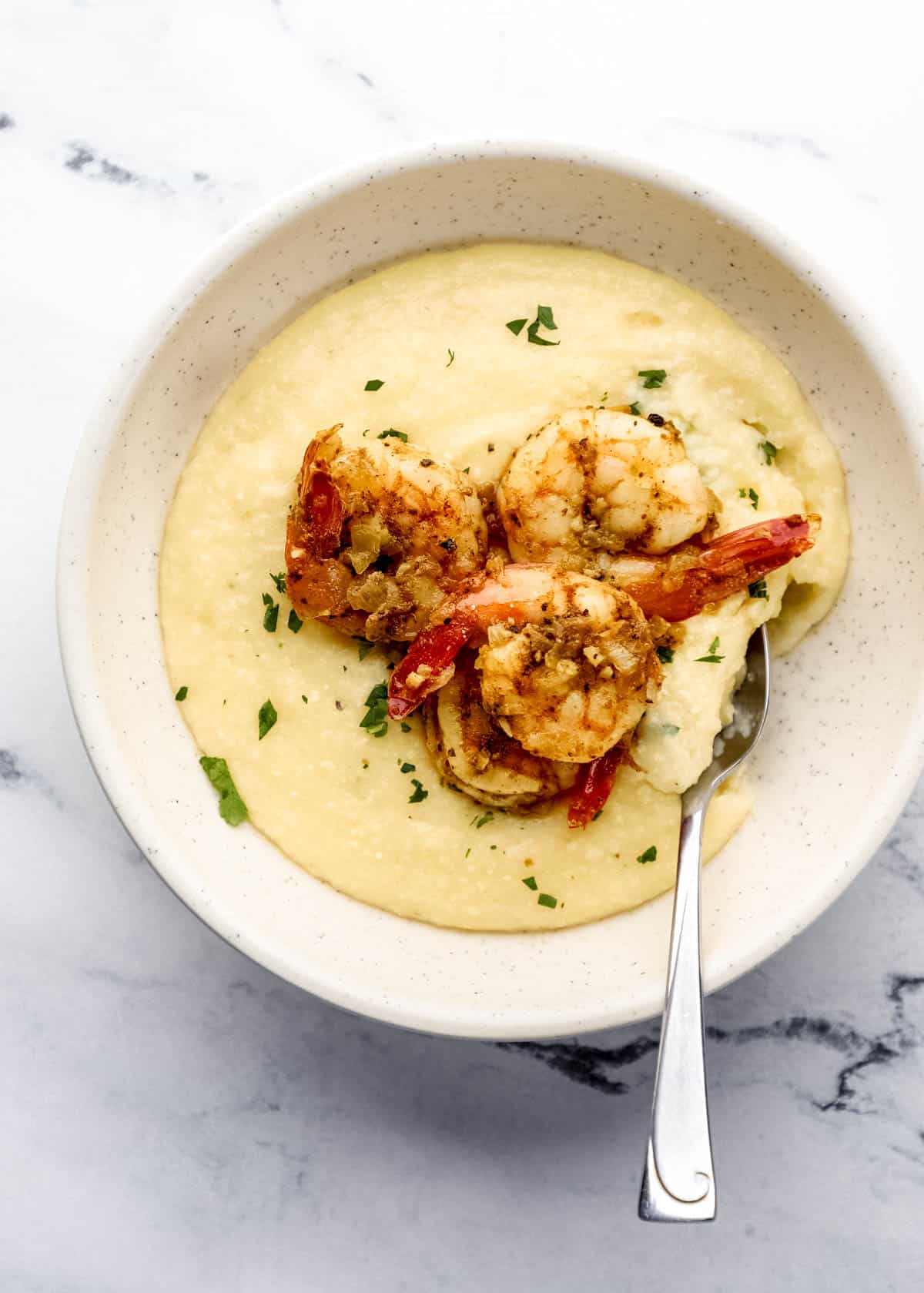 Bowl of grits topped with shrimp with spoon in it on marble surface. 