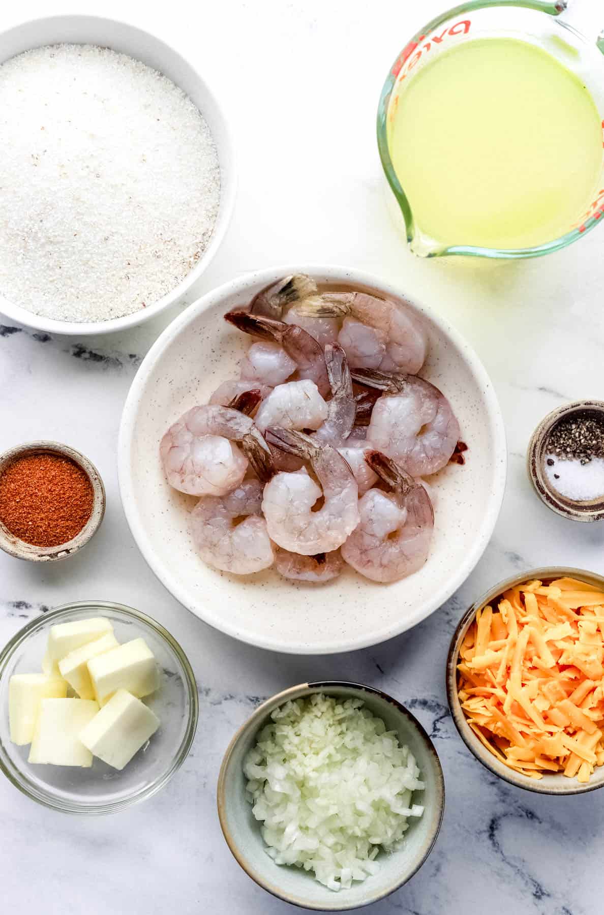 Overhead view of ingredients needed to make shrimp and grits in separate bowls on marble surface. 