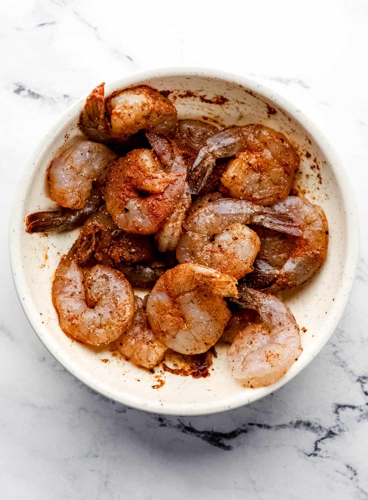 Old Bay seasoned raw shrimp in large white bowl on marble surface. 