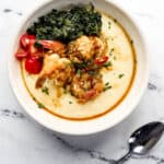 Overhead view of white bowl with shrimp and grits in it by a spoon on marble surface.