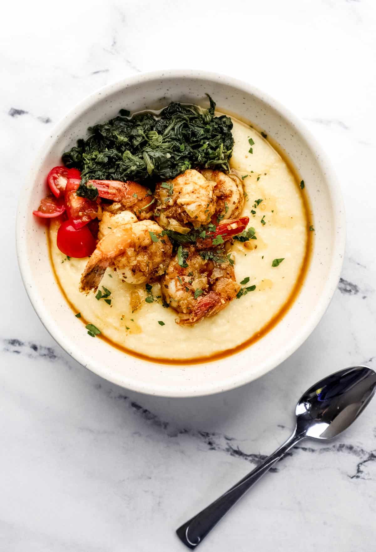 Overhead view of shrimp and grits in a white bowl by a spoon on marble surface. 