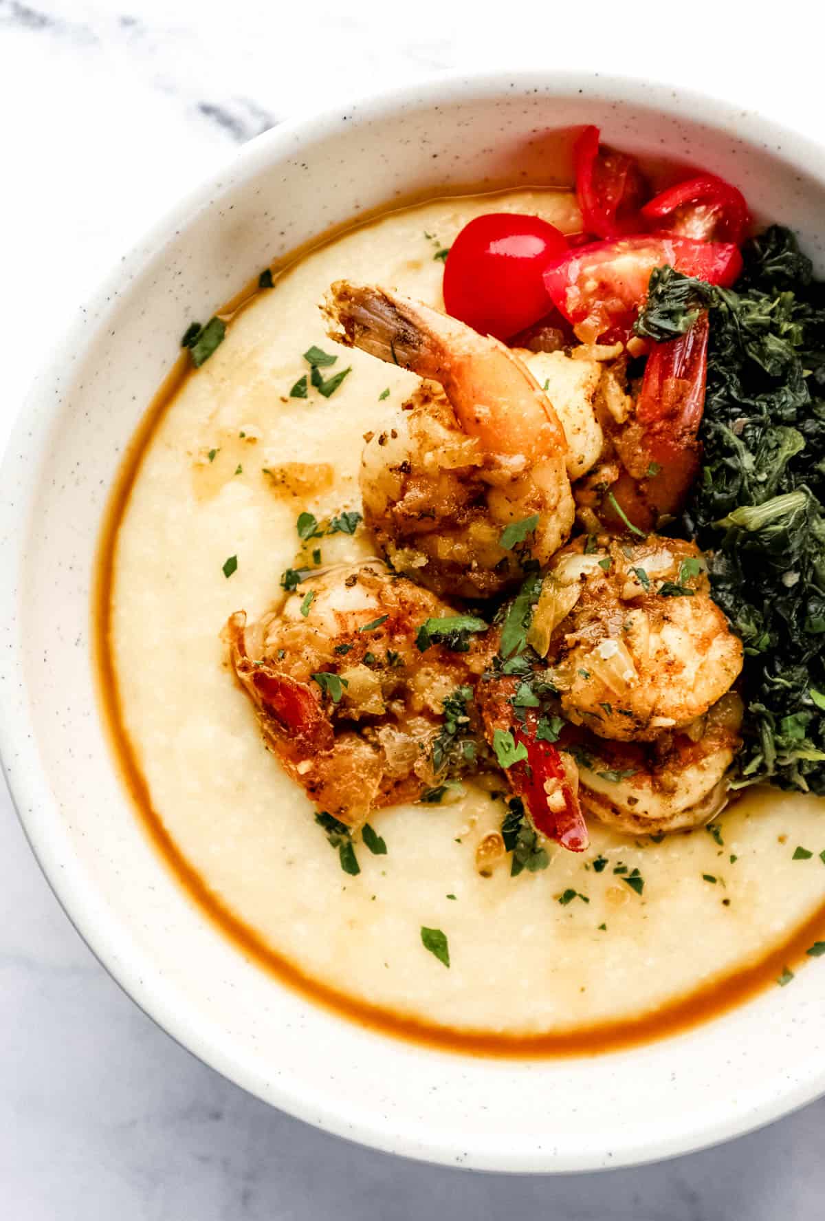 Overhead view of bowl of shrimp and grits topped with spinach and tomatoes on marble surface. 