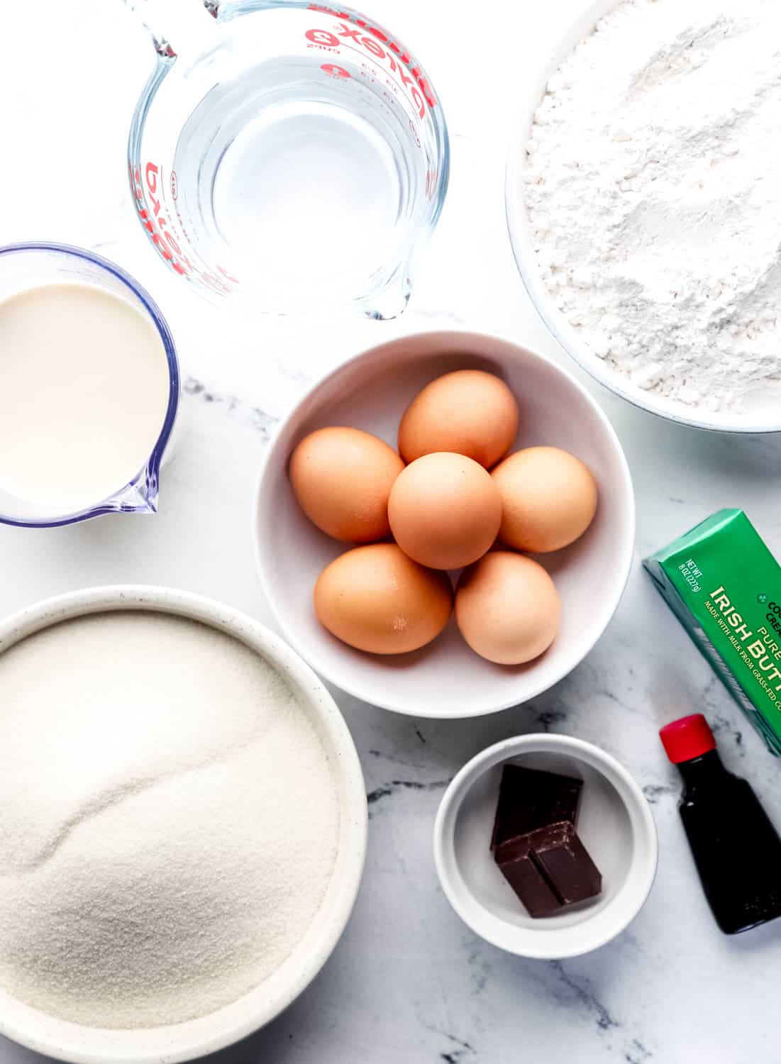 Overhead view of ingredients needed to make chocolate cake in separate bowls and containers on marble surface. 