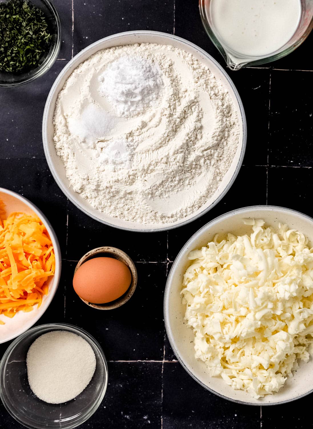 Overhead view of ingredients needed to make biscuits in separate bowls on black tile surface. 