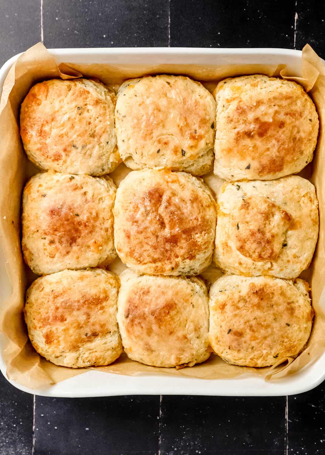 Overhead view of finished biscuits in square parchment lined baking dish on black tile surface. 