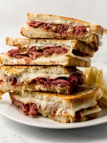Close up side view of corned beef grilled cheese sandwiches stacked on white plate with chips on it.