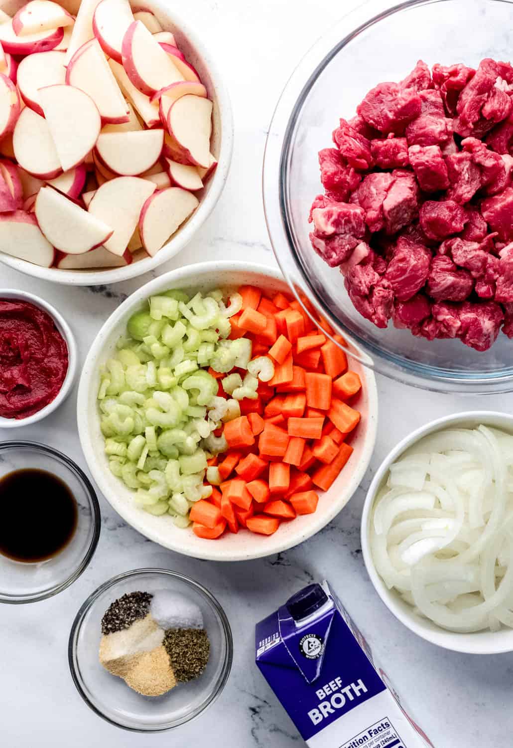 Overhead view of ingredients needed to make beef stew in separate bowls on marble surface. 