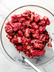 Overhead view of seasoned beef with tongs in glass mixing bowl.