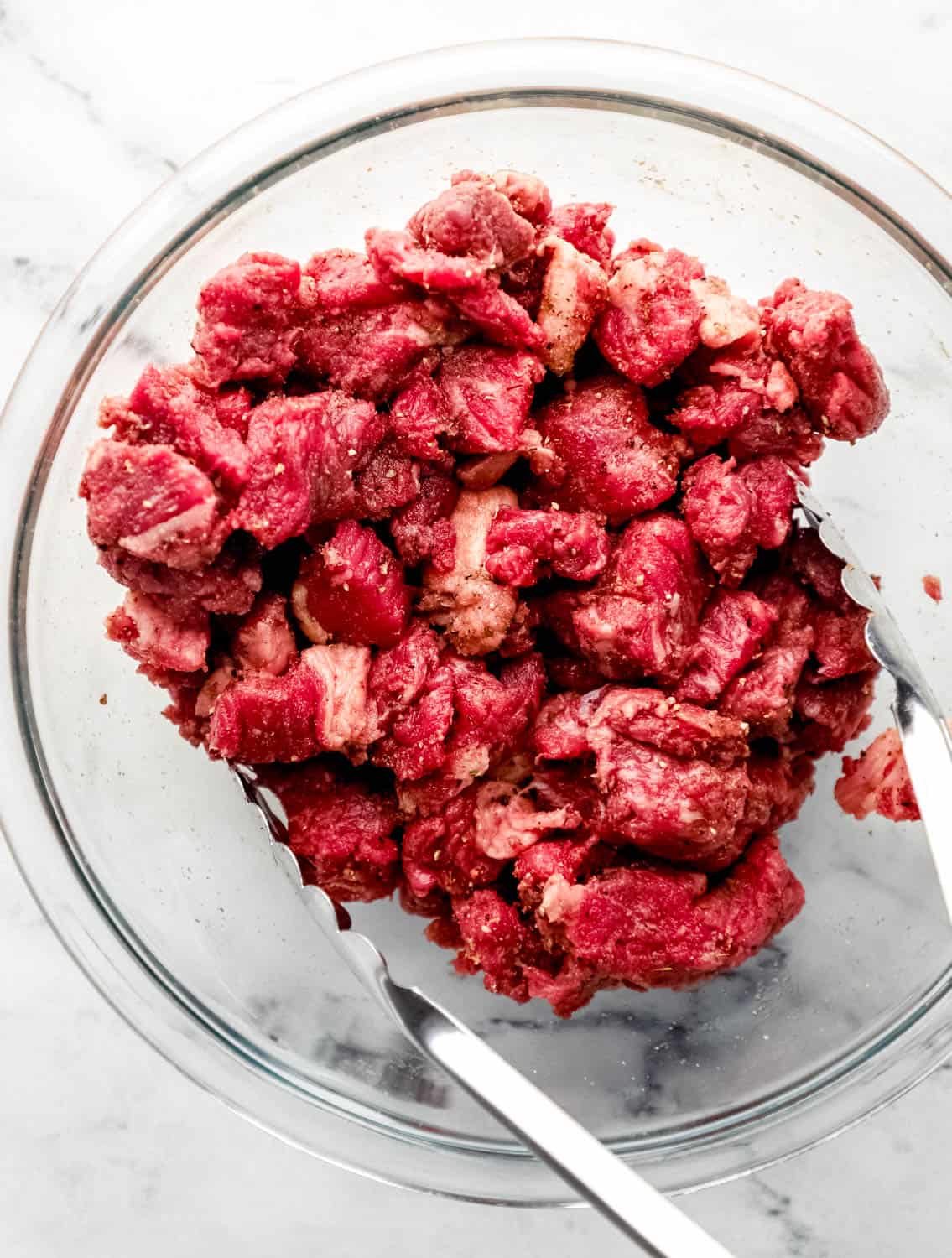 Seasoned raw beef in large glass mixing bowl with tongs in it on marble surface. 