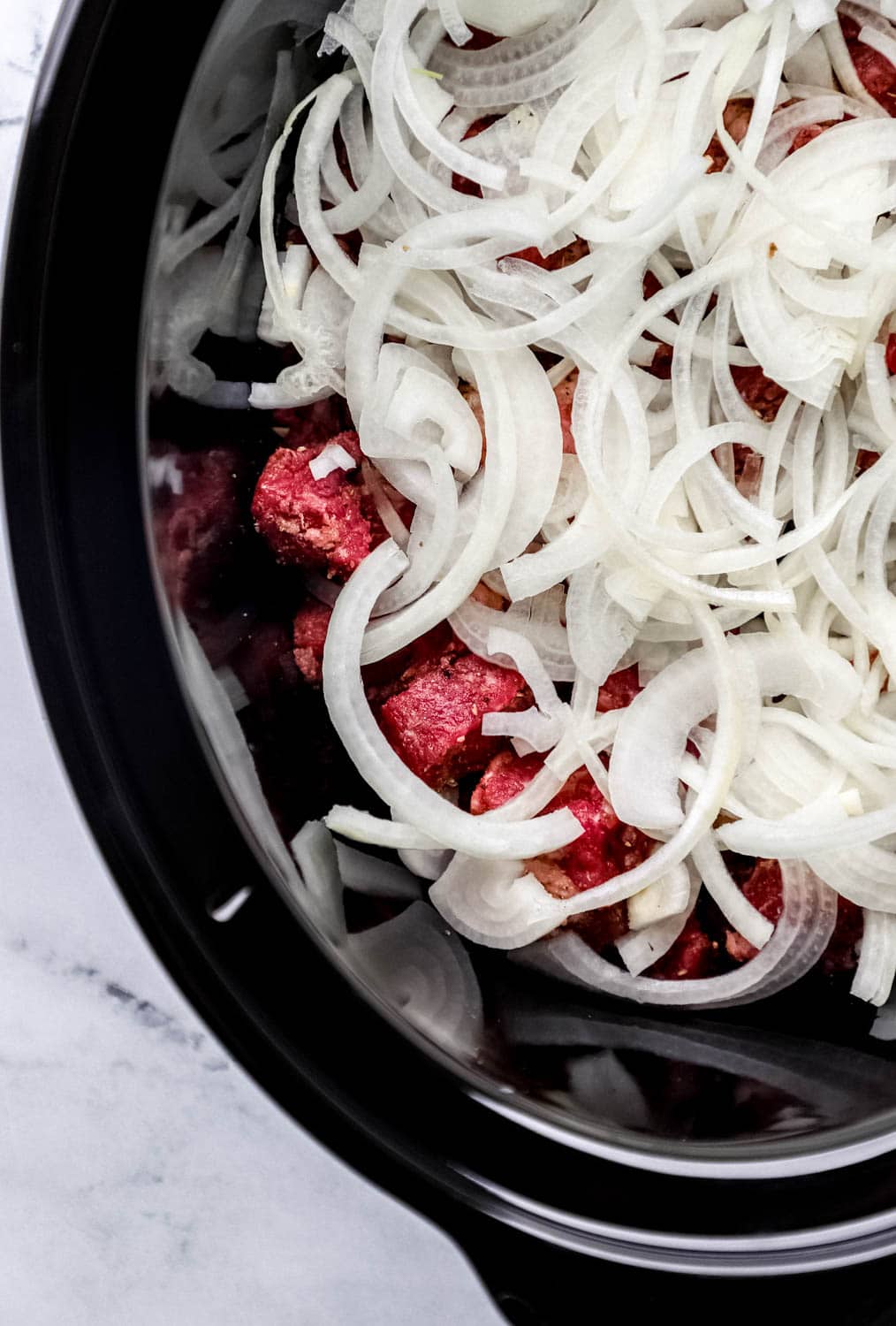 Overhead view of uncooked seasoned beef and sliced onion added to crock pot. 