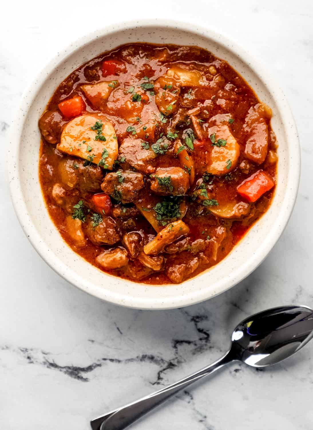 Overhead view of white bowl with beef stew in it on marble surface by a spoon. 