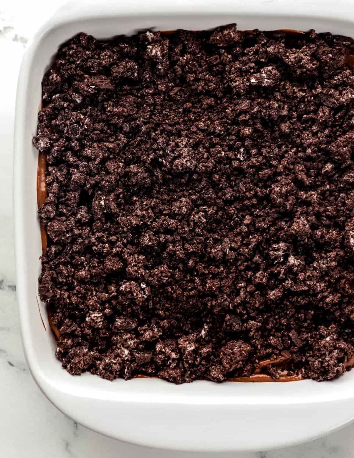 Crushed oreo cookies added on top of chocolate pudding in white baking dish. 