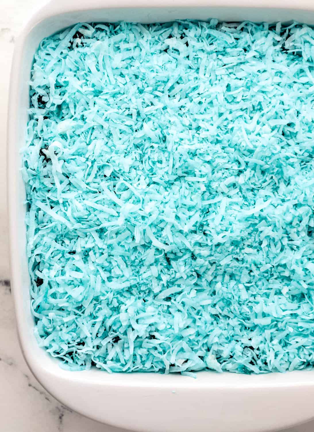 Colored shredded coconut added to the top of the crushed cookies in the white baking dish. 