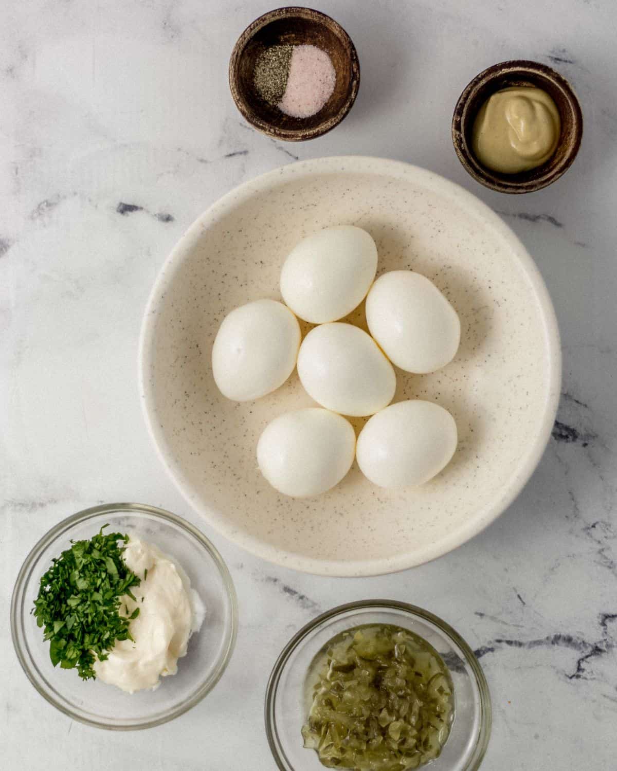 Overhead view of ingredients needed to make egg salad in separate bowls on marble surface. 