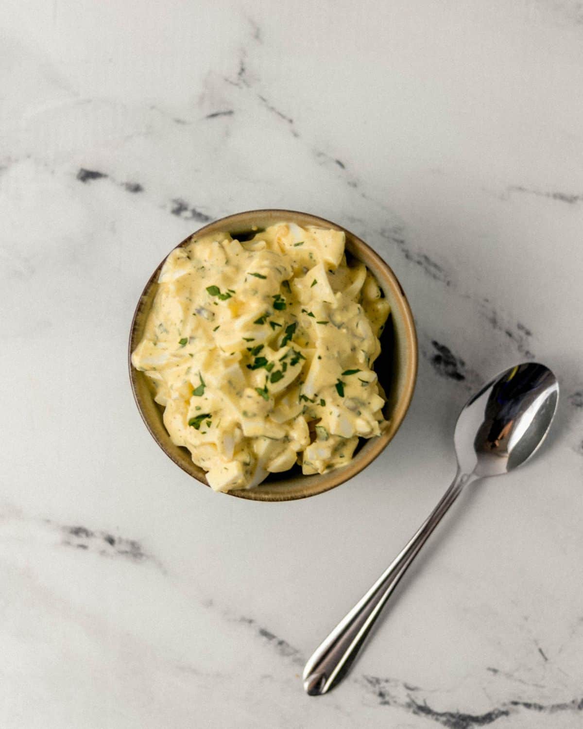 Small bowl with egg salad in it beside a spoon on marble surface. 