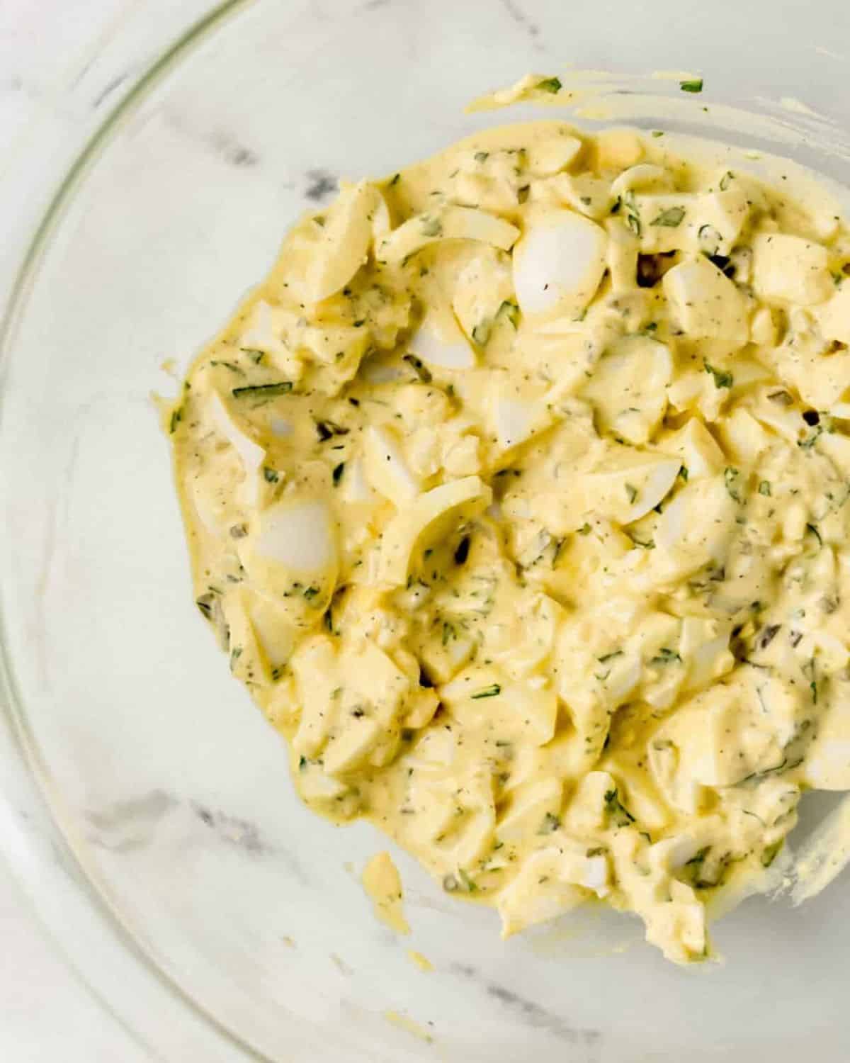 Egg salad ingredients combined in large glass mixing bowl on marble surface. 
