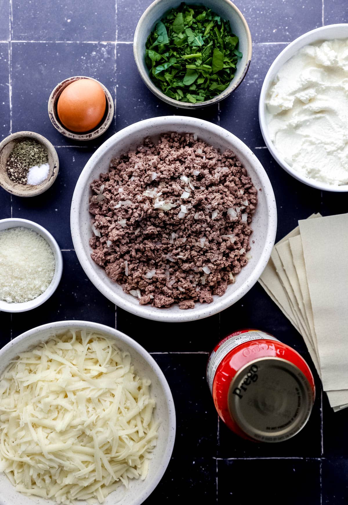 Overhead view of ingredients needed to make lasagna in separate bowls and containers on black tile surface. 