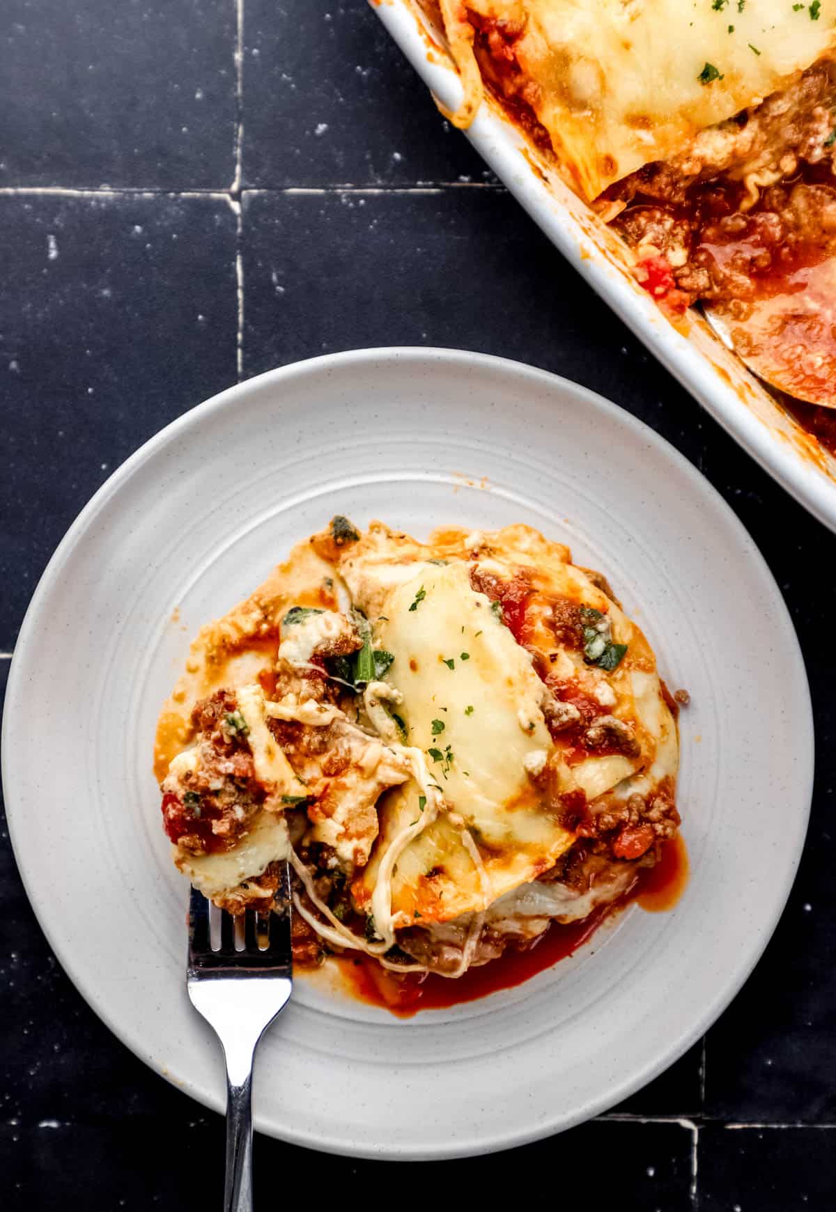 Overhead view of white plate with serving of lasagna on it with the rest of the pan in the background on black tile surface. 