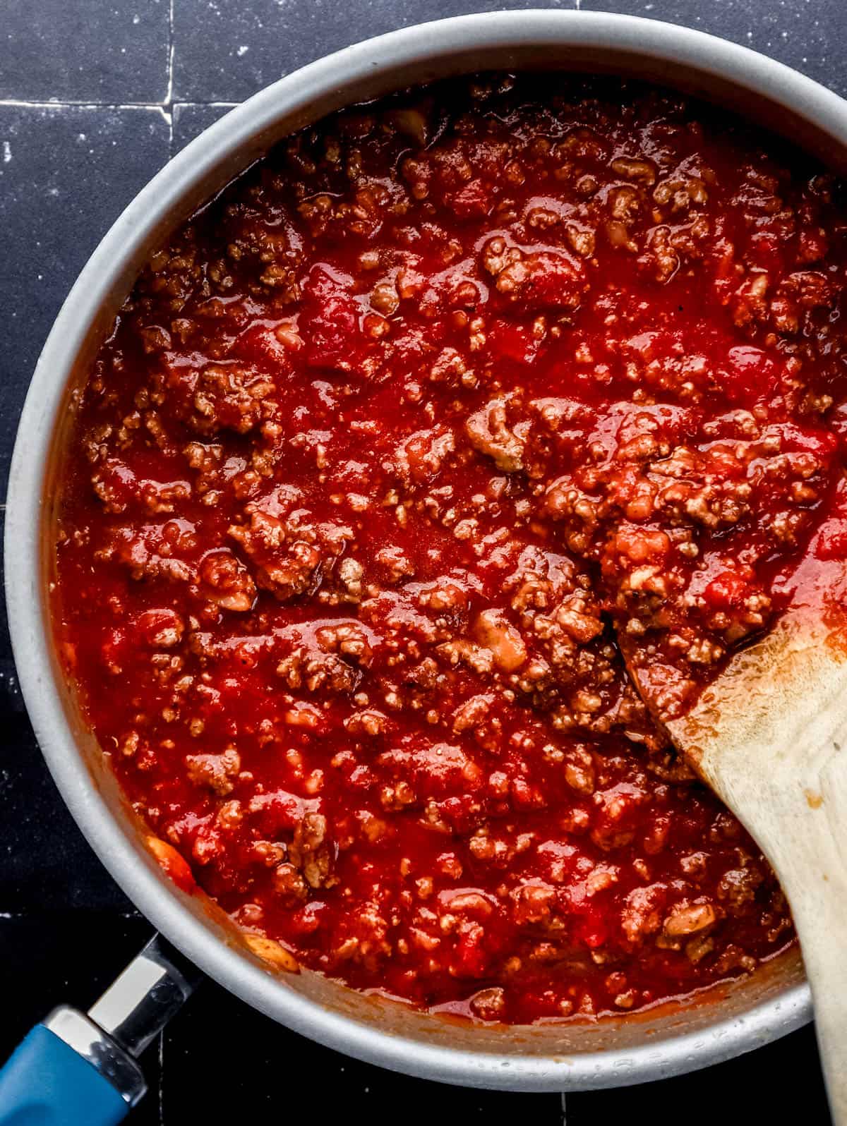 Overhead view of meat sauce ingredients in large skillet with wooden spoon. 
