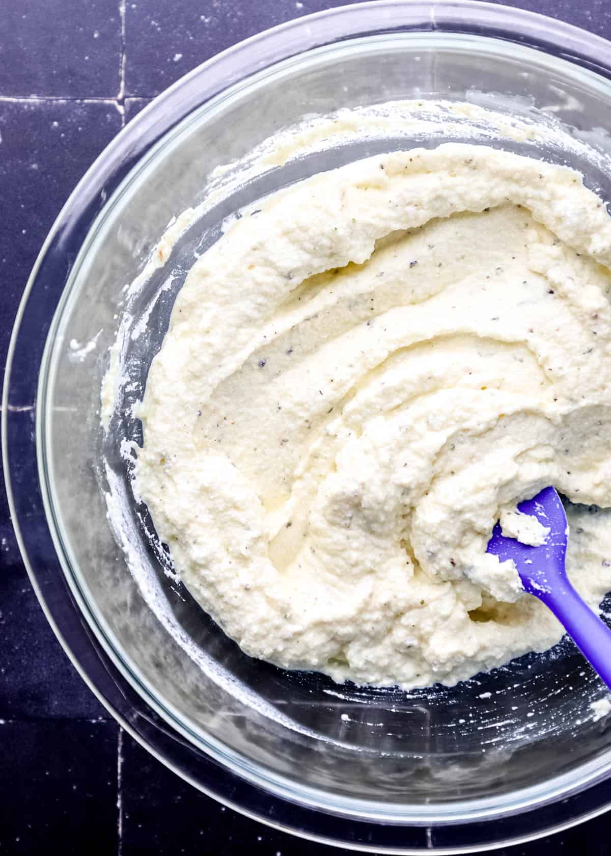 Ricotta cheese mixture in large glass mixing bowl with blue rubber spatula in it. 