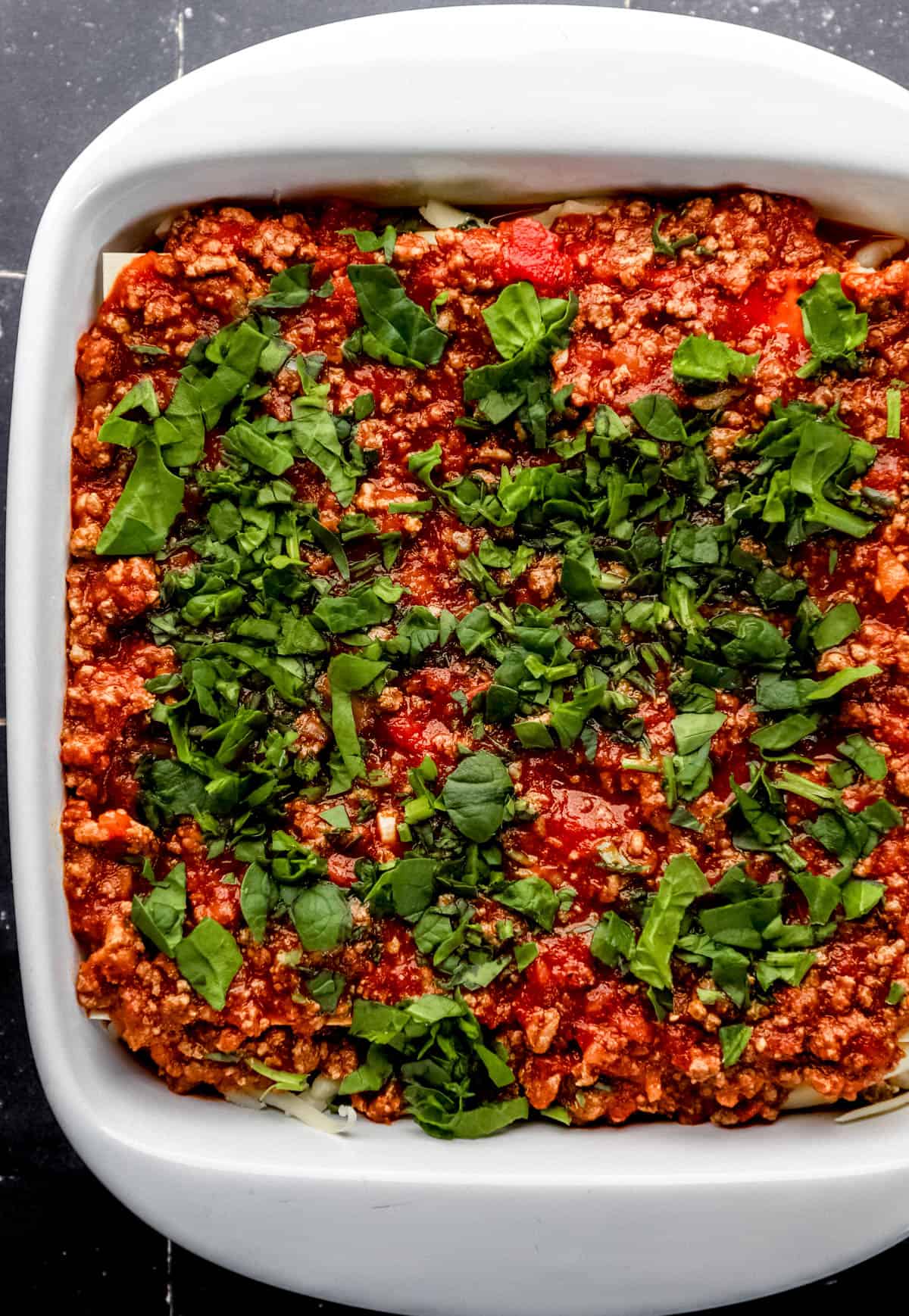 Overhead view of second layer of lasagna starting with meat sauce and spinach in white baking dish. 