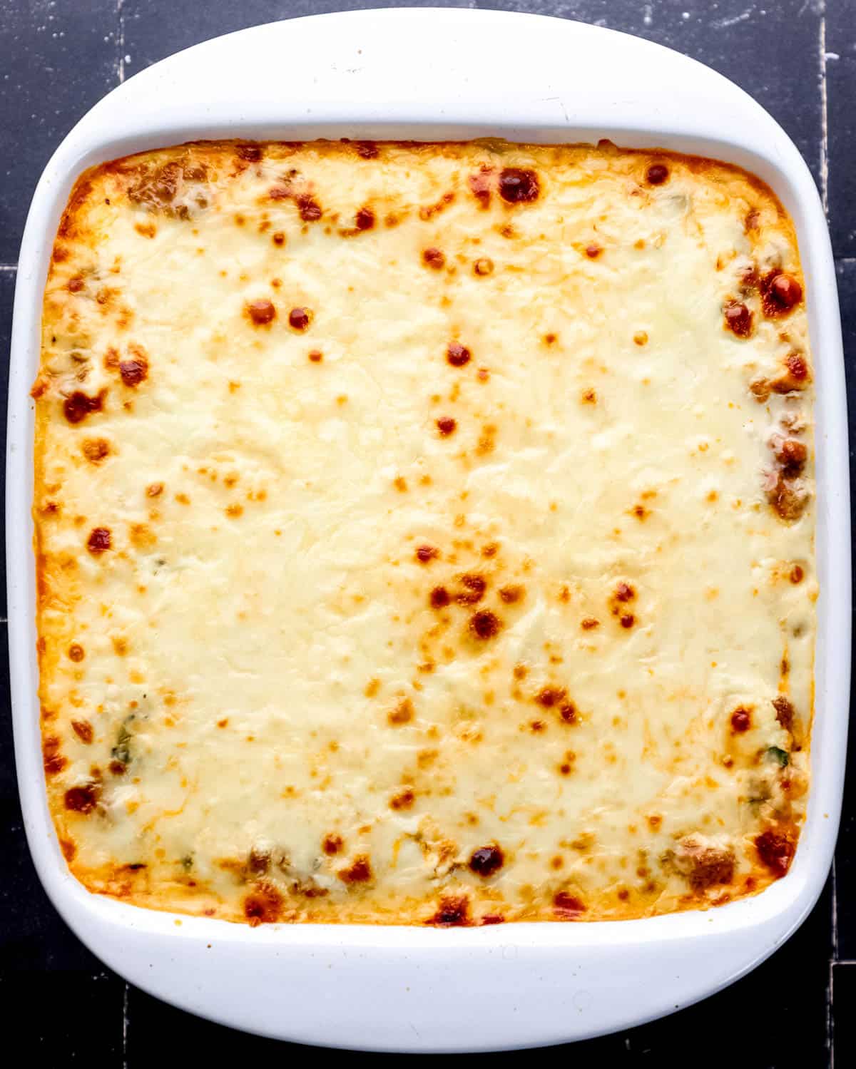 Overhead view of baked lasagna in square white baking dish on black tile surface. 