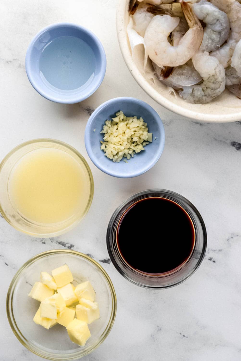 Overhead view of ingredients needed to make shrimp in separate bowls on marble surface. 