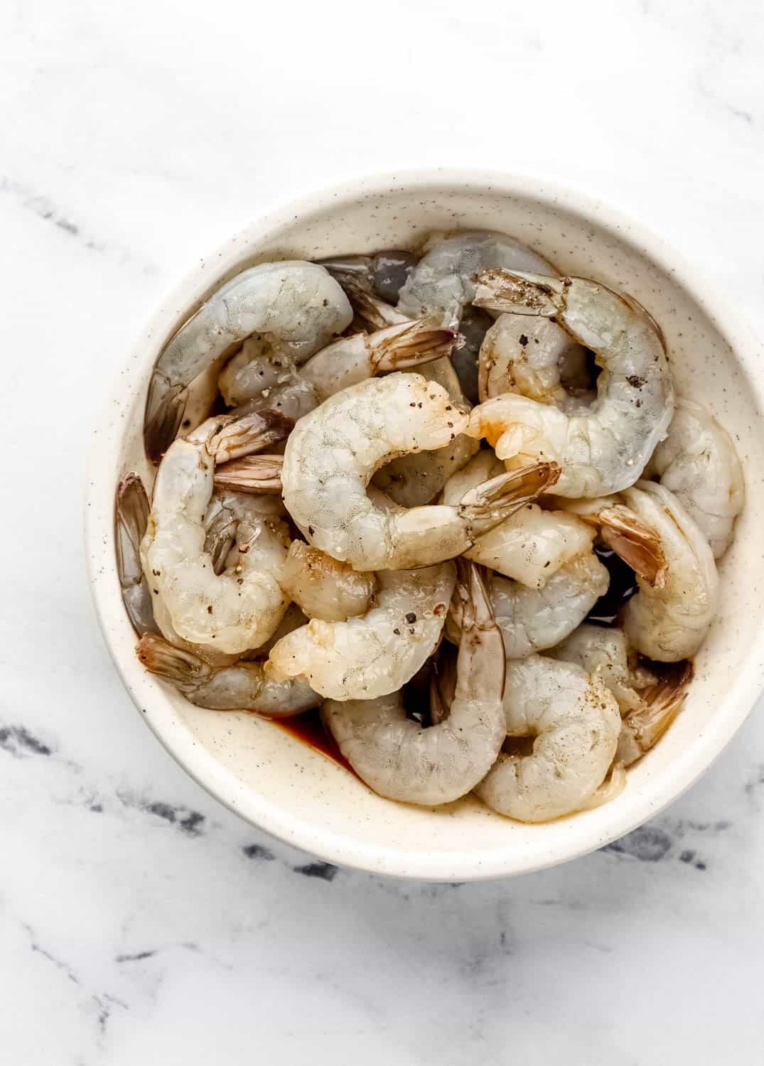 Overhead view of seasoned and marinated raw shrimp in white bowl on marble surface. 