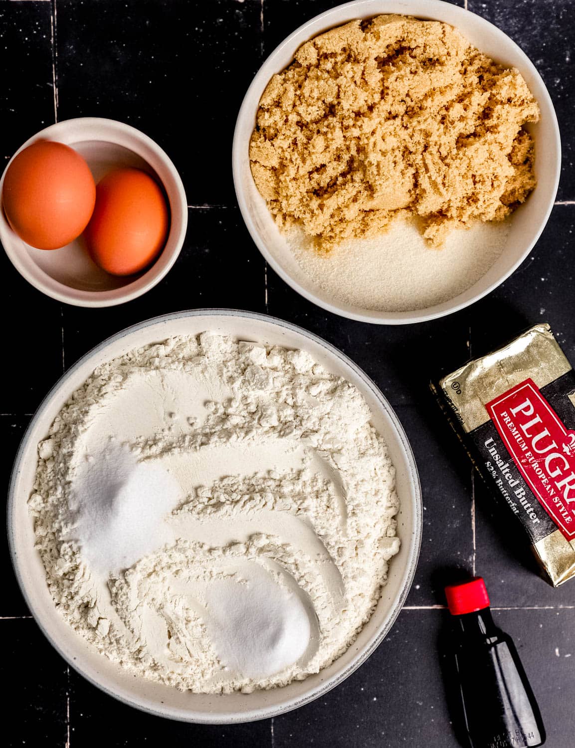 Overhead view of ingredients needed to make cookies in separate bowls on black tile surface. 