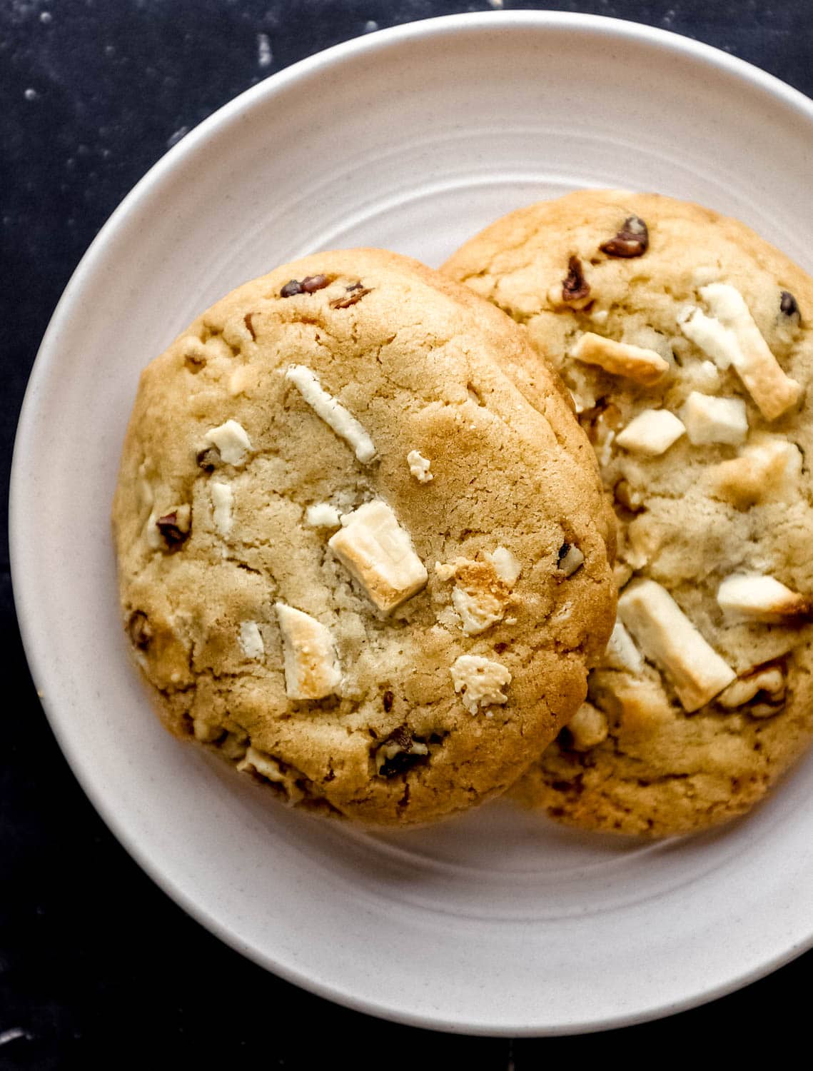 Overhead view of two baked cookies on white plate on top of black tile surface. 