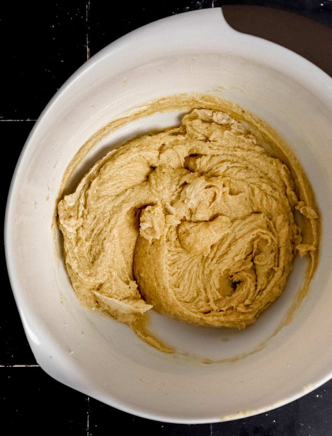 Overhead view of wet cookie dough ingredients combined in large white mixing bowl on black tile surface. 