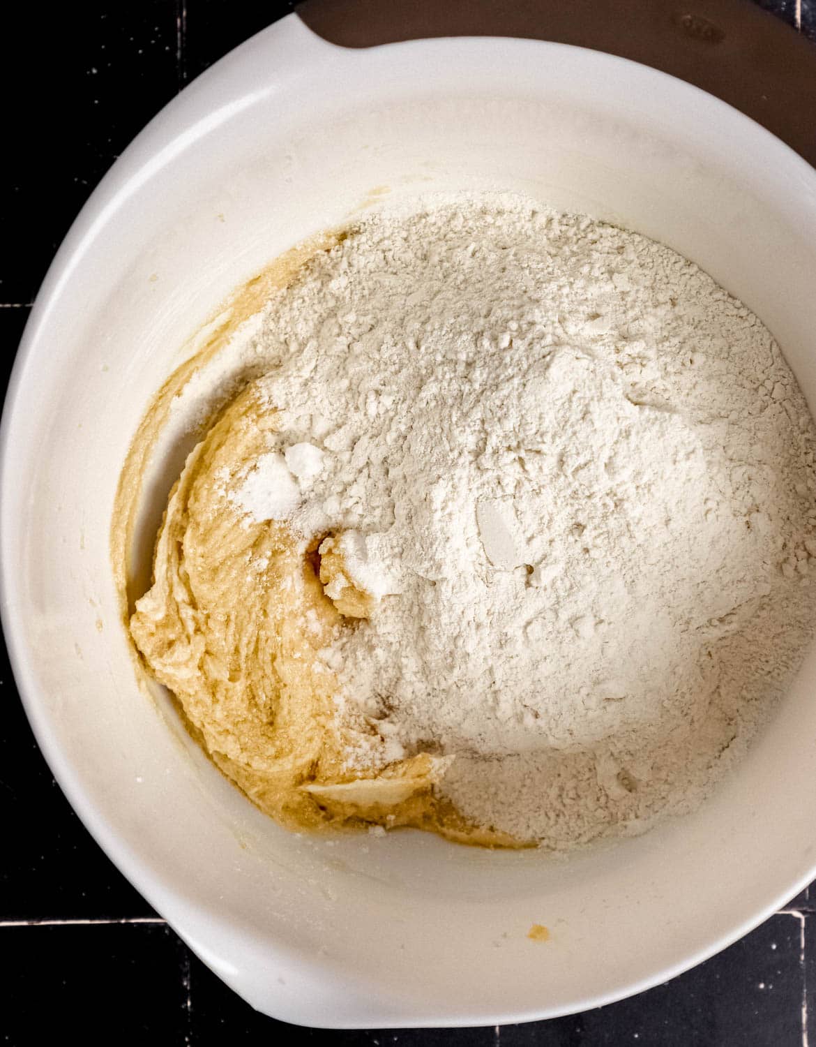 Overhead view of dry ingredients added to wet cookie ingredients in large white mixing bowl. 