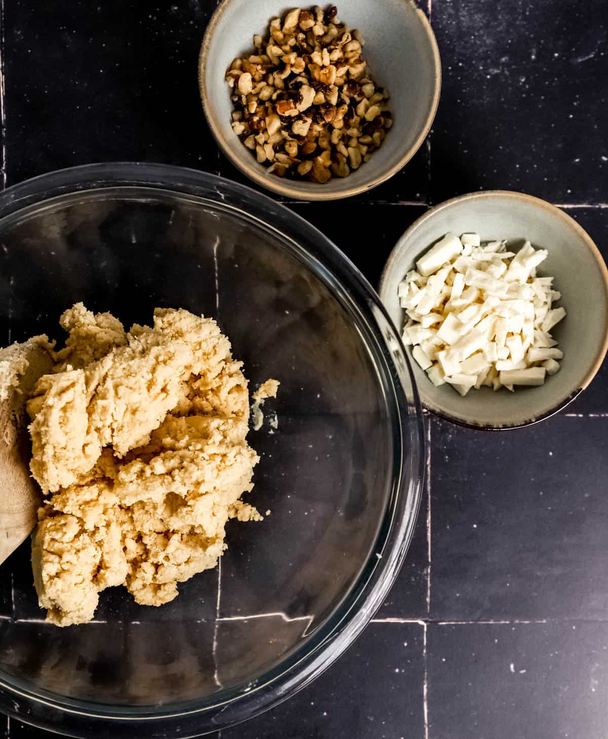 Overhead view of bowl of cookie dough beside two small bowls, one with nuts and the other with white chocolate. 