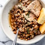 Overhead view of white bowl with black eyed peas, pork, corn bread, and spoon in it.