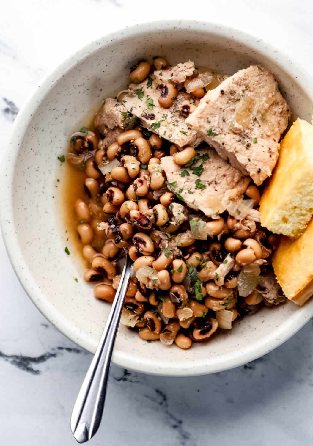 Bowl with black eyed peas, pork chops, corn bread, and spoon in it. 