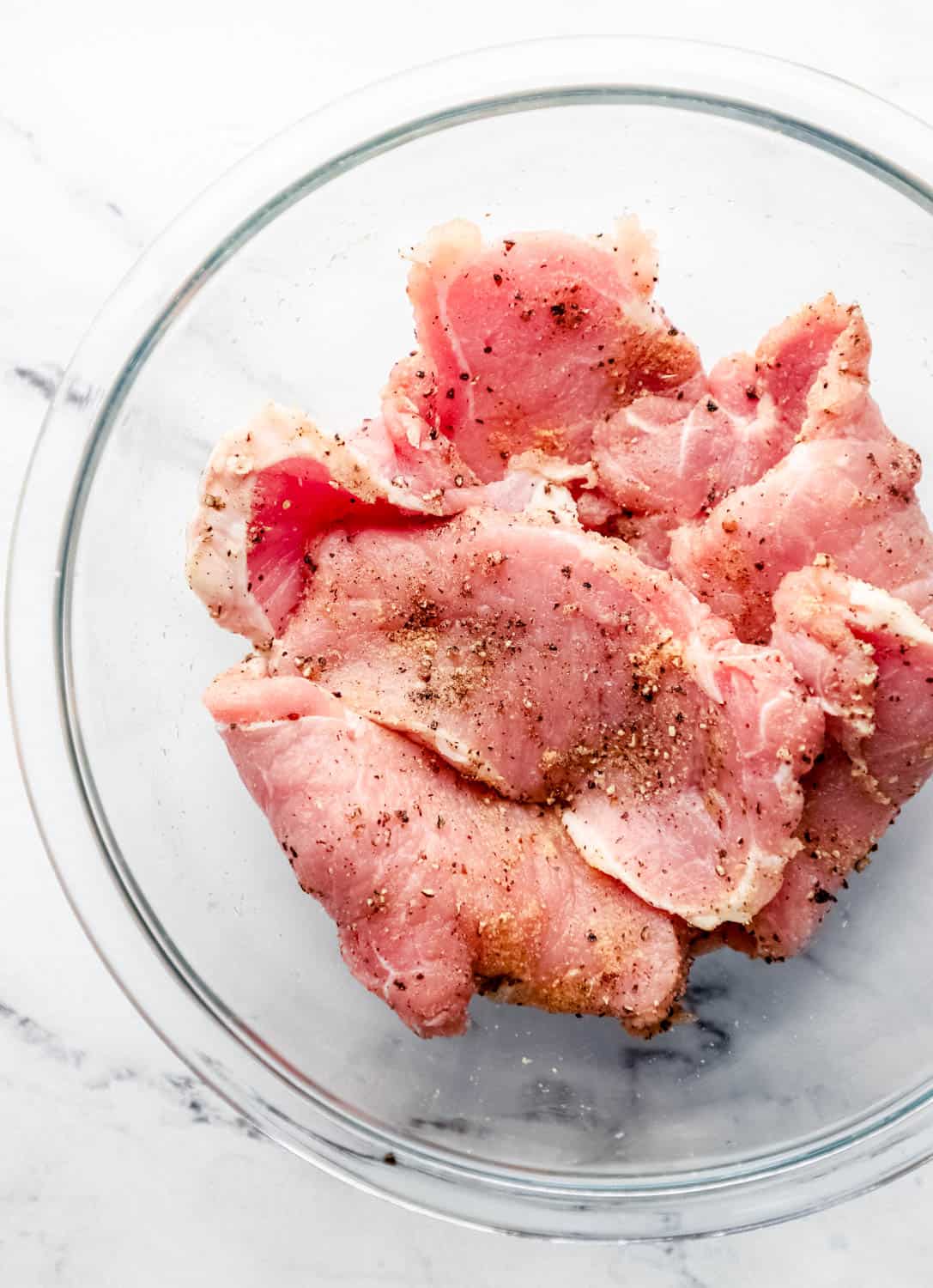 Overhead view of large glass bowl with seasoned pork chops in it on marble surface. 