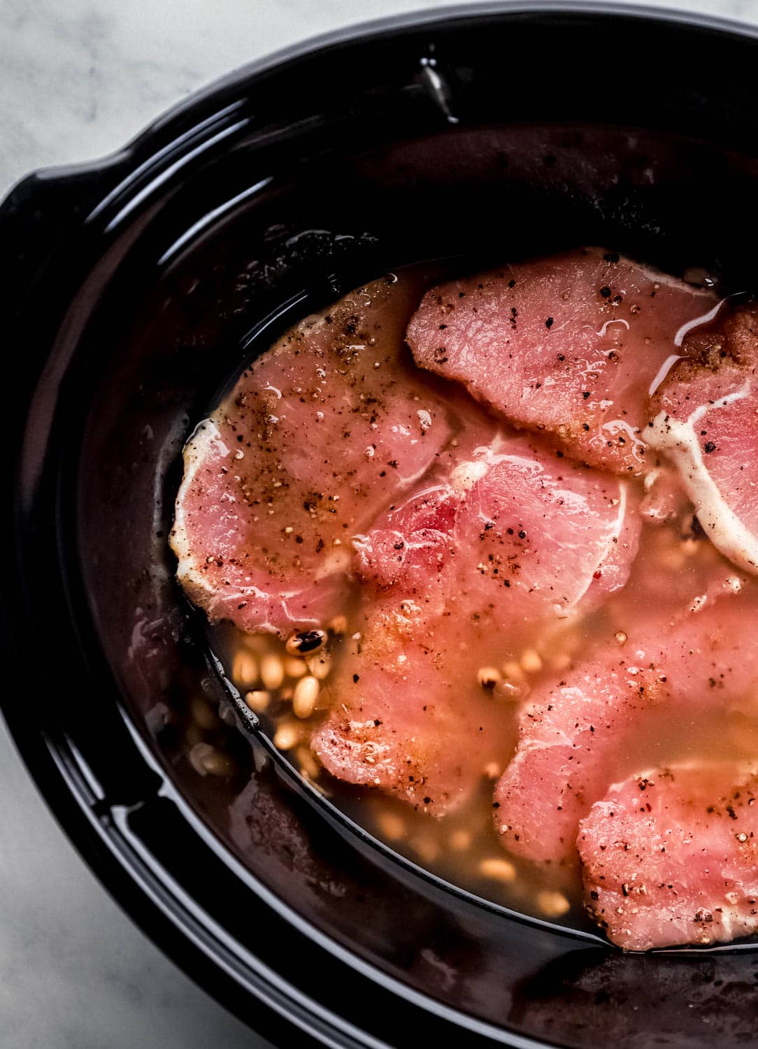 Overhead view of slow cooker with canned peas and seasoned pork chops in it. 