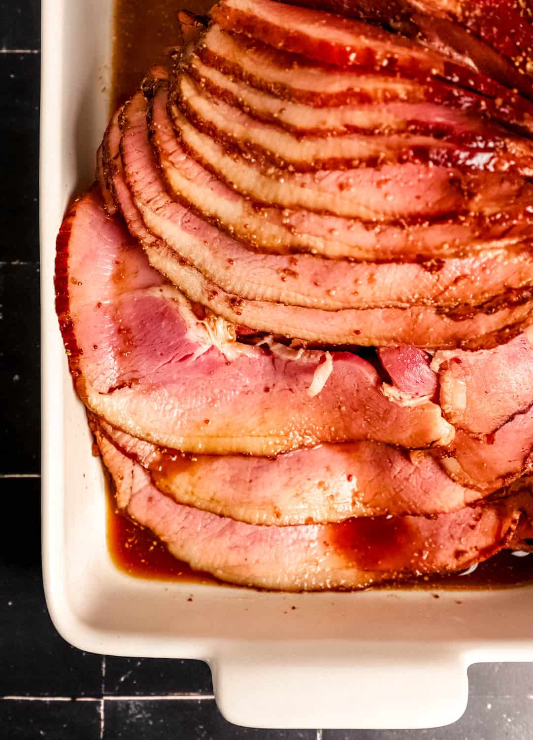 Overhead view of sliced baked ham in roasting pan on black tile surface.