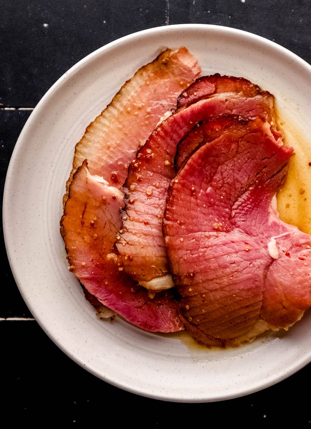 Overhead view of slices of ham on white plate with glaze on black tile surface. 