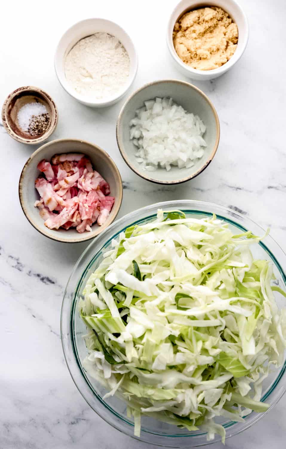 Overhead view of ingredients needed to make cabbage in separate bowls on marble surface. 
