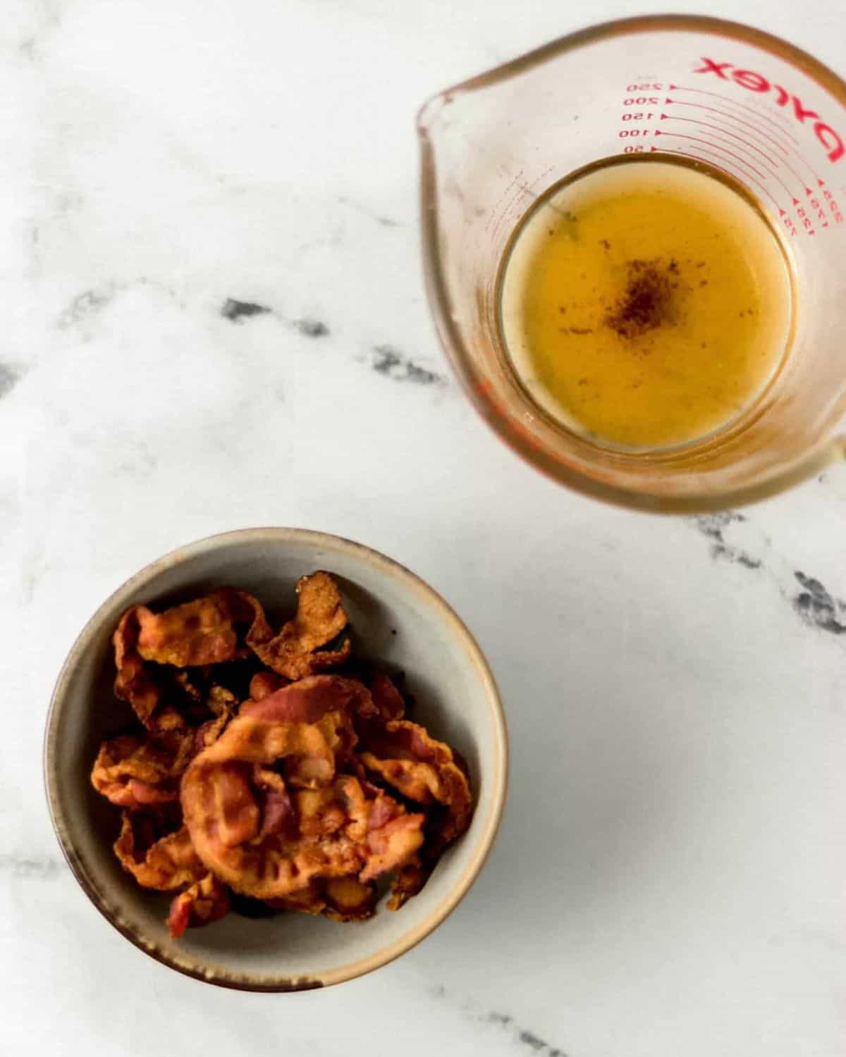 Overhead view of cooked bacon in a small bowl and measuring cup with bacon grease in it on marble surface. 