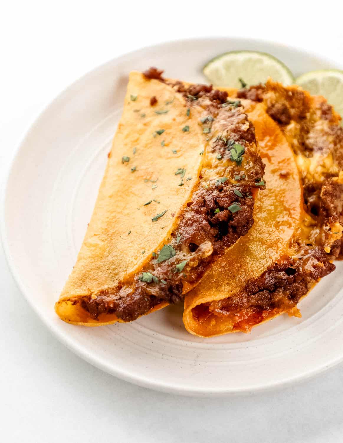 Close up view of two baked tacos on white plate with slices of lime on white surface. 