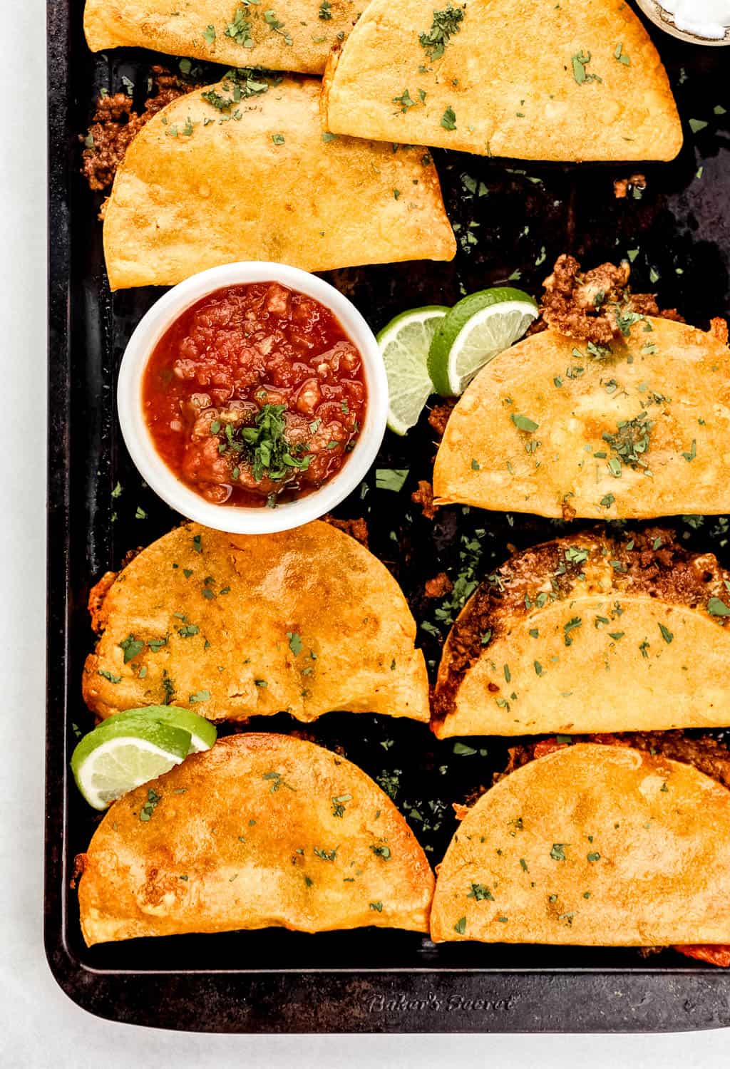 Overhead view of baked tacos on sheet pan with salsa and slices of lime. 