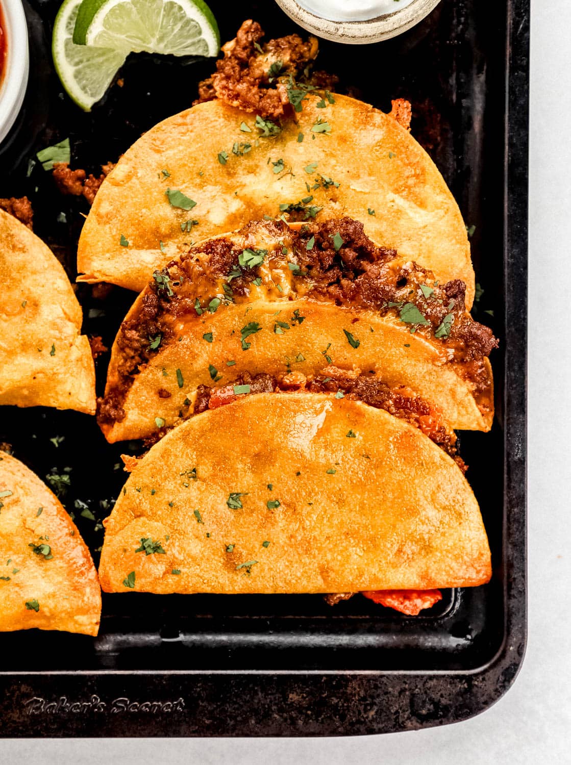 Finished tacos on baking sheet with slices of lime and small bowls of salsa and sour cream. 